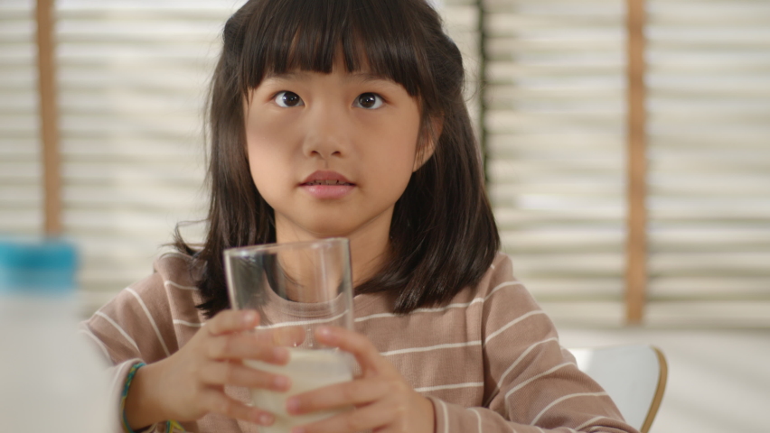 Slow motion - Portrait of Cute Asian Little Girl Drinking milk sits in the kitchen. Cute Girl having breakfast and enjoying natural fresh milk in the morning at home. Healthy and Natural Food. Royalty-Free Stock Footage #1058928863