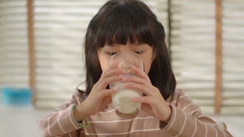 Slow motion - Portrait of Cute Asian Little Girl Drinking milk sits in the kitchen. Cute Girl having breakfast and enjoying natural fresh milk in the morning at home. Healthy and Natural Food.