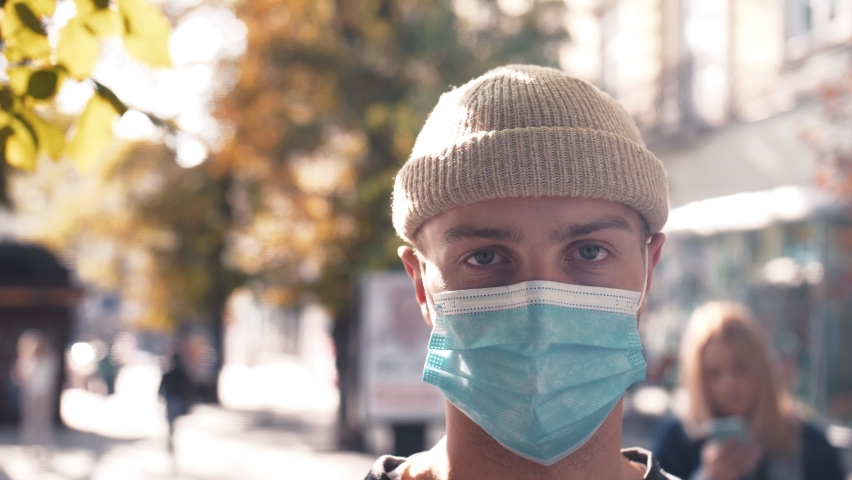 Close up of face of young caucasian handsome man in medical mask looking straight to camera. Portrait of man at street. Coronavirus concept.