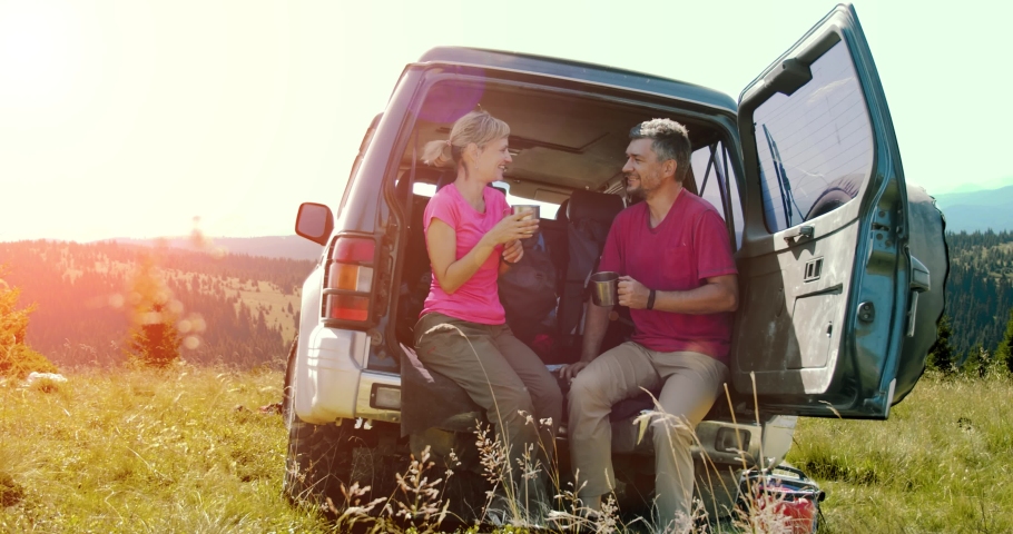 Young man and woman eholding cups with drinks and sitting near the car in the mountain during sunny day.