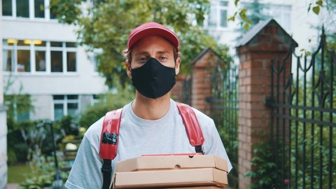 Portrait of young courier delivery man holding pizza in carton boxes, wearing protective face mask, waiting for customer in residential area. Delivery services.