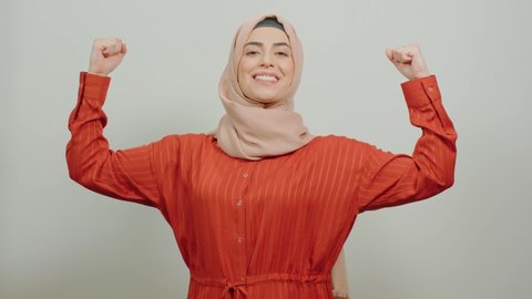 Young muslim woman in turban, posing on white background, smiling broadly for the camera while showing her strong biceps. Strong woman portrait.