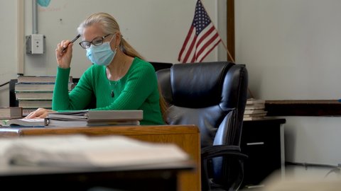Teacher in school working at desk wearing medical face mask with sanitizer on desk with PPE and US flag behind.