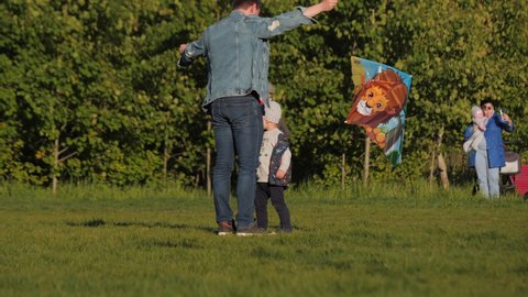 KAZAN, TATARSTAN/RUSSIA - MAY 28 2020: Young father flies colorful kite spending time with little son on lush grass in green spring park on sunny day on May 28 in Kazan