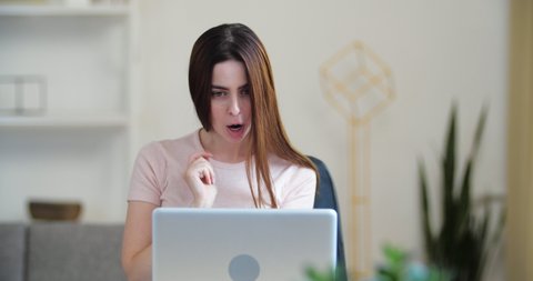 Successful student young woman sits in front of laptop, communicates on webcam online, reads good news, happily shakes her head and hair, victorious movements, makes gesture like shoots at screen
