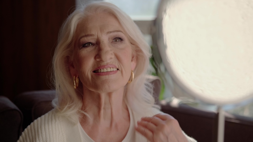 Beautiful well-groomed mature woman with gray hair, with jewelry looks in the mirror and smiles. An adult pensioner with a beautiful face and long hair is taking care of herself. Senior woman portrait