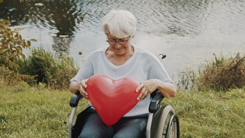 Senior woman showing heart shaped balloon near the river. Love and care concept. High quality 4k footage