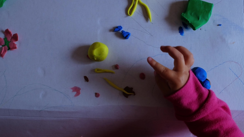 From above view of unrecognizable little child playing with colorful modelling clay on white table at home