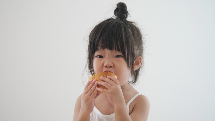 Asian preschool toddler girl in cute top knot hair eating homemade bread deliciously with both hands. Wearing white tank top underwear. Isolated in white background. Safety and healthy food ingrediant. Royalty-Free Stock Footage #1058944391