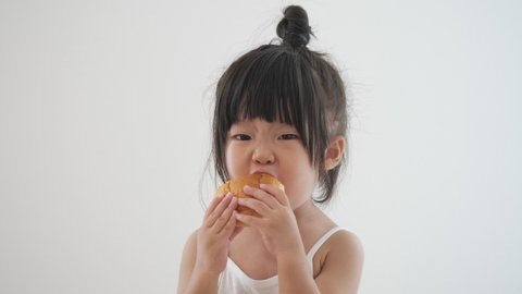 Asian preschool toddler girl in cute top knot hair eating homemade bread deliciously with both hands. Wearing white tank top underwear. Isolated in white background. Safety and healthy food ingrediant.