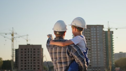 Father and son happy Family concept. Little boy and his dad engineer builder architect with safety helmets, on background new buildings and construction cranes on site. Future profession, 4 K slow-mo