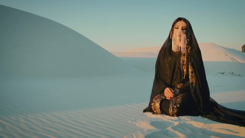 A beautiful young woman in a black oriental dress sits in a desert. head is covered with a scarf, face is hidden by golden veil mask. Pretty girl in image of an Egyptian Bedouin, gold accessories
