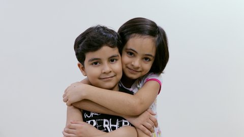 A beautiful little sister coming and hugging her younger brother with a smile . Smiling young brother is hugged by his sister from back when she sees him posing in front of the camera