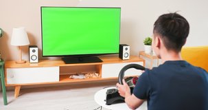 Asian gamer boy is playing a racing game by steering wheel with green chroma key TV screen at home