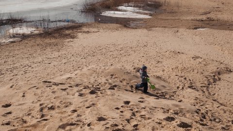 A boy in a gray jacket, hat, boots and blue pants runs down the sand hill, holds a green childrens shovel and a bucket in his hands, sunny weather, a beach by the river