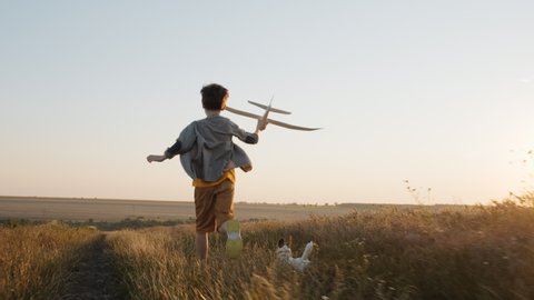 Happy Boy Runs Wheat field  holding airplane in his hand, runs with dog on meadow against sunset of summer day towards bright sun slow motion lens flares. Child lifestyle. Travel. Childhood. Pet