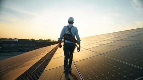 Maintenance assistance technical worker in uniform is checking an operation and efficiency performance of photovoltaic solar panels on roof at sunset. Sun clean green energy