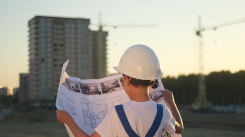 Little boy child future engineer builder architect with safety helmets with drawing, looking on new buildings and construction cranes on site. Dreaming of future profession. Dream concept, 4 K slow-mo