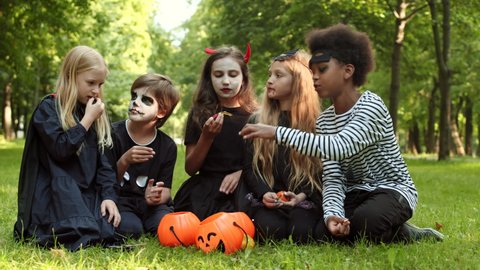 Wide shot of five multiethnic kids wearing different Halloween costumes are sitting on grass in park, taking candies and lollipops out of pumpkin buckets and eating them