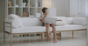 little boy is sitting on couch at home and using video call for communicating with friends by laptop, self-isolation