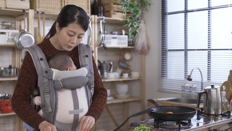 Japanese female carrying a baby in the front is turning food in heated pan cautiously. and moving egg with a spatula slowly in order not to burn her daughter with hot oil splatter.