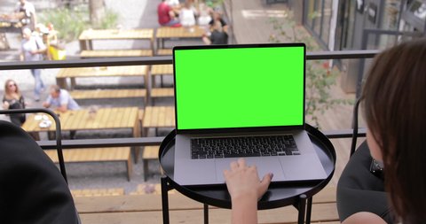 Young Woman at Coffee Shop Working on a Laptop MacBook Pro with Green Mock-up Screen. She's Sitting On a Bean Bag Chair in Coworking open space outdoor office. Over the Shoulder Camera Shot.