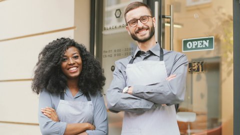 Caucasian barista man and african american barista woman standing at entrance to coffee shop. Beautiful people in aprons and gray shirts standing outside and smiling with their arms crossed.
