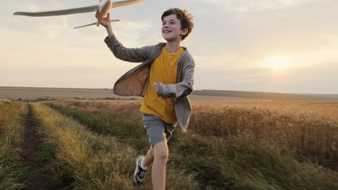 Happy boy runs of Wheat Field  holding airplane in his hand run on summer meadow of sunset of summer day towards bright sun slow motion. Child plays lifestyle. Childhood. Agro Farm. Go Everywhere