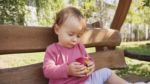Baby child girl eat bite ripe apple fruit sitting on a bench in apple tree garden