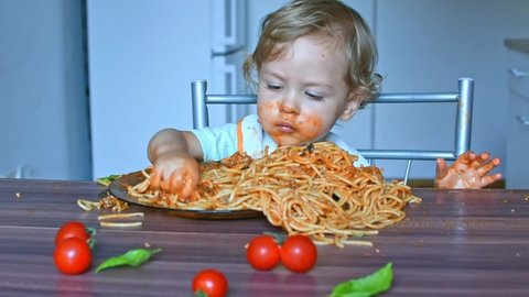 Happy sweet baby boy making a mess in the kitchen while eating spaghetti. Funny toddler child with home made pasta and tomato sauce