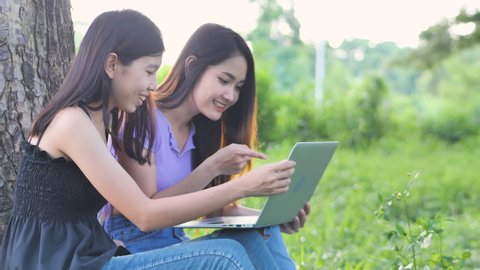 Two young lady sitting near tree at the park, they are look at the laptop, feel having fun laugh and happy together on holiday. Online video call concept. Technology in life concept. Slow motion. 
