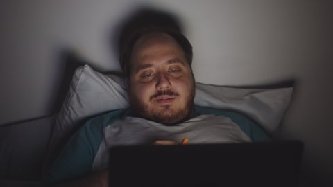 Fat guy on diet watching video on laptop at home in bed at night eating carrot sticks. Stout man enjoying movie on laptop in bedroom eating healthy snack late at night