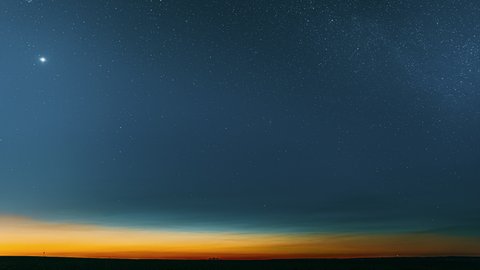Night Starry Sky With Glowing Stars Above Countryside Field Landscape In Early Spring. Bright Glow Of Planet Venus In Sky Among The Milky Way Galaxy Stars. Sky In Lights Of Sunset Dawn. 4K Timelapse.