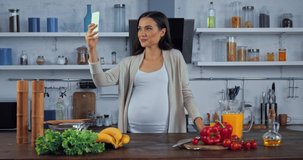Pregnant woman taking selfie with smartphone near vegetables in kitchen