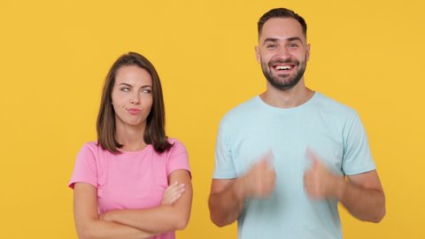 Young fun couple friends bearded man woman 20s in basic t-shirts posing isolated on yellow background studio. People emotions lifestyle concept. Look camera showing thumbs up down different gestures