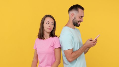 Young couple friends bearded man spy woman in basic t-shirt standing back to back isolated on yellow background studio. People emotion lifestyle concept Look at gadget using mobile cell phone chatting