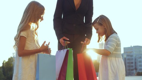 Successful mom shows gifts to two little daughters standing on street threesome, close up. Happy faces of children girls looking at opening shopping bag. Family bought new birthday presents in store