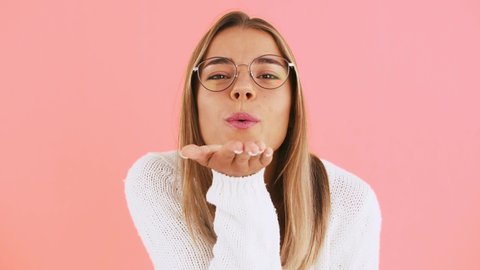 Young woman is winking, sending you an air kiss and smiling while posing against pink studio background