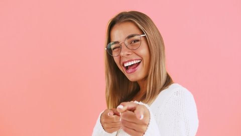 Young lady in glasses and white jumper is pointing at you by forefingers, winking and laughing posing on pink background