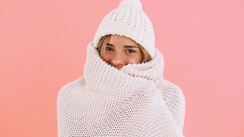Woman in sweater and hat is feeling cold. She wrapping herself in warm cover and smiling posing on pink background