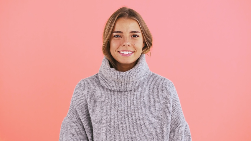 Woman is covering her face with neck of sweater, looking around, winking and laughing while posing on pink background | Shutterstock HD Video #1058984630
