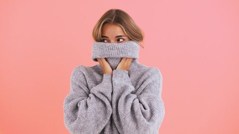 Woman is covering her face with neck of sweater, looking around, winking and laughing while posing on pink background