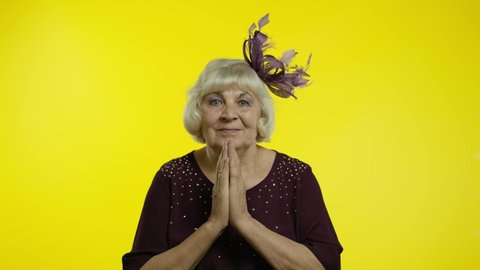 Please, I am begging. Positive senior old blond woman appealing to camera, keeping prayer gesture and asking help, apologize. Elderly stylish lady grandma on yellow background in studio. 6k downscale