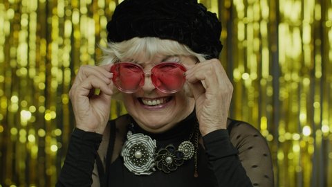 Lovely coquettish mature old woman touching red sunglasses and winking playfully, flirting and blinking eye as having some interesting idea, cheering up with happy look. Elderly grandma. 6k downscale