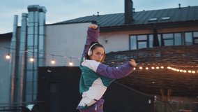 Dynamic caucasian woman freestyle artist wearing sportive overalls and headphones listening to music enjoying rhythm gesturing and breakdancing on the roof.