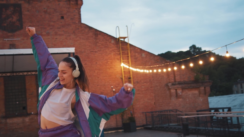 Dynamic caucasian woman freestyle artist wearing sportive overalls and headphones listening to music enjoying rhythm gesturing and breakdancing on the roof. Royalty-Free Stock Footage #1058987828