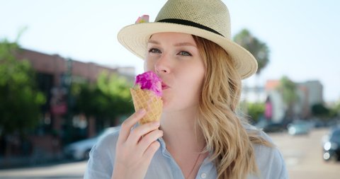 Smiling blonde woman eating tasty ice cream in waffle cone, she is seeing friend, waving hand and smiling inviting to join her in the cafe. Woman with delicious Italian gelato ice cream on summer day
