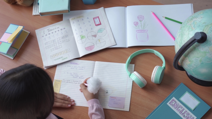 Top above view of indian latin preteen primary school girl student, child pupil studying at home sitting at desk writing, doing homework, learning at table with exercise books and supplies. Royalty-Free Stock Footage #1058989070
