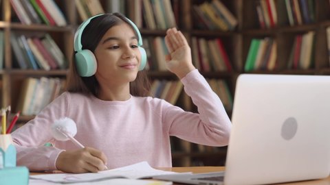 Happy indian latin preteen girl school pupil wearing headphones raising hand distance learning online at virtual lesson class with teacher tutor on laptop by video conference call studying at home.
