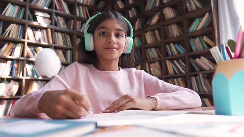 Cute indian latin preteen girl school kid child pupil wearing headphones raising hand distance learning online talking to camera at virtual lesson class by video conference call at home, webcam view.